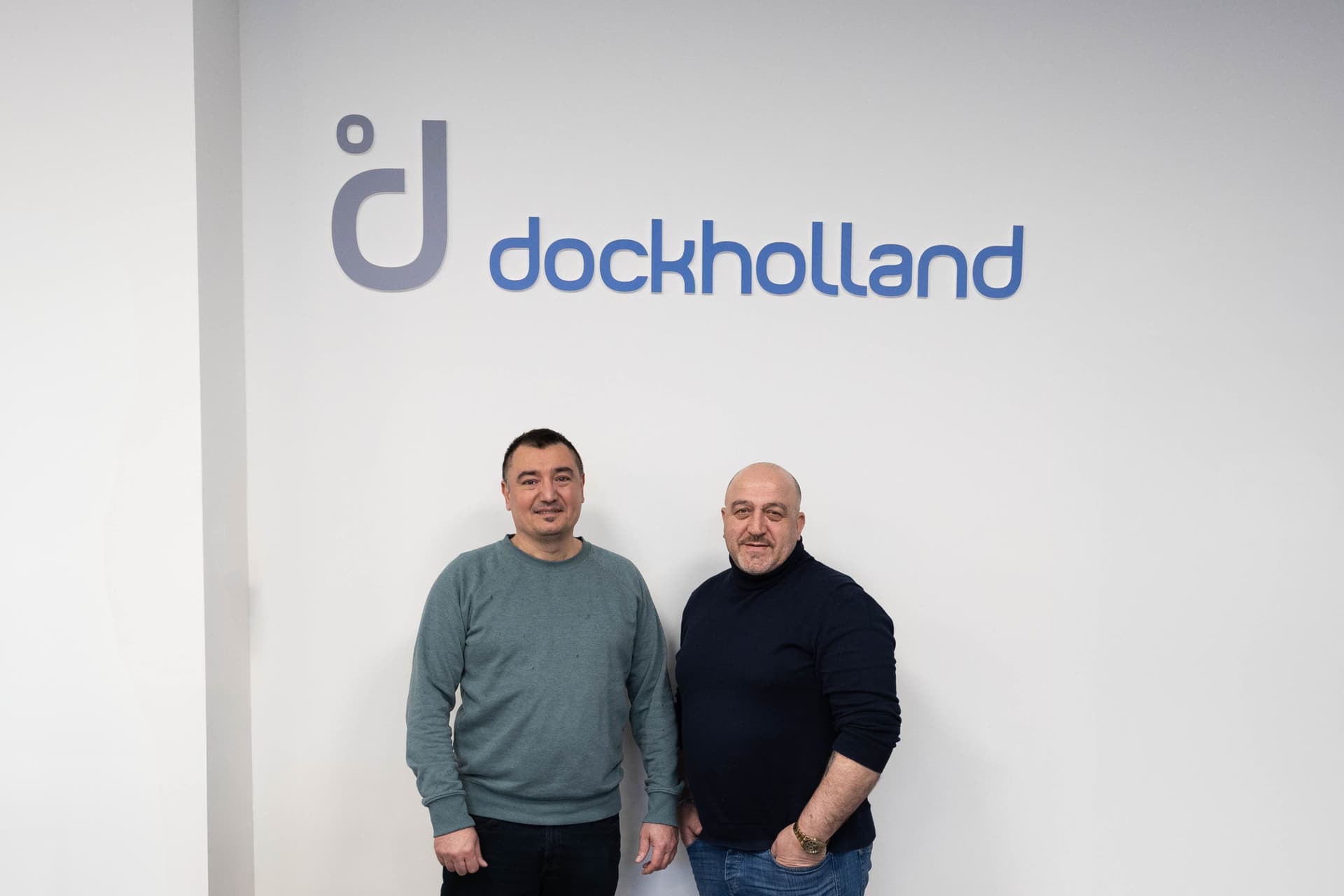 Directeur en medewerker poseren naast elkaar voor een witte muur met het logo van DockHolland op de achtergrond